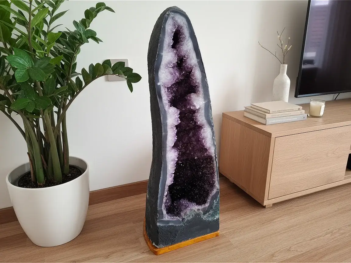 Velika ametistna geoda z globoko vijoličastimi kristali, postavljena na lesenih tleh ob moderni hrastovi komodi, belem keramičnem loncu z zelenim rastjem ter minimalističnih dekorativnih dodatkih, ki poudarjajo razkošen interier.