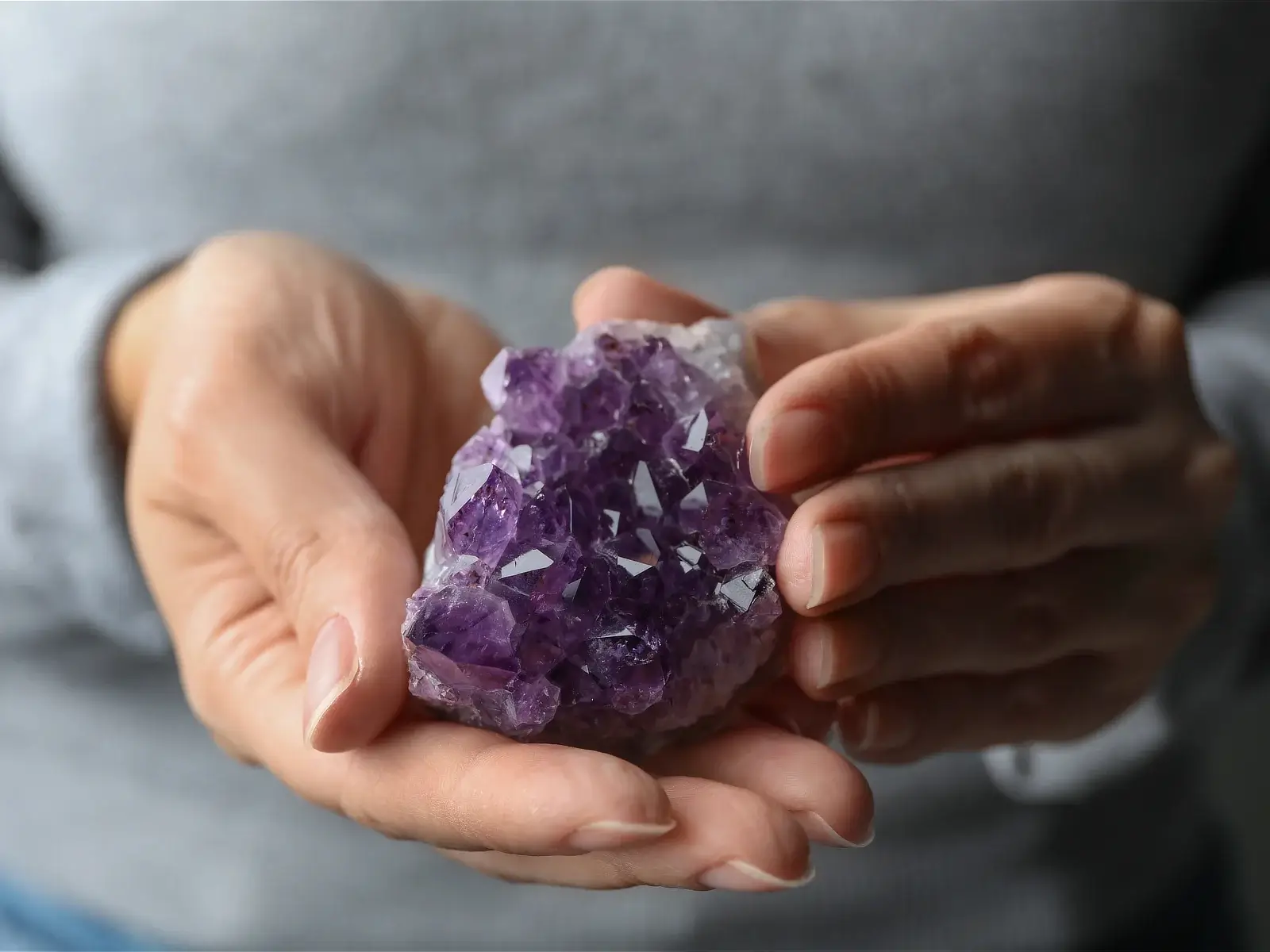 Roke nežno držijo naravni grozd ametistnih kristalov z globoko vijoličnimi kristalnimi tvorbami, jasno izoblikovanimi fasetami in naravno mineralno strukturo.