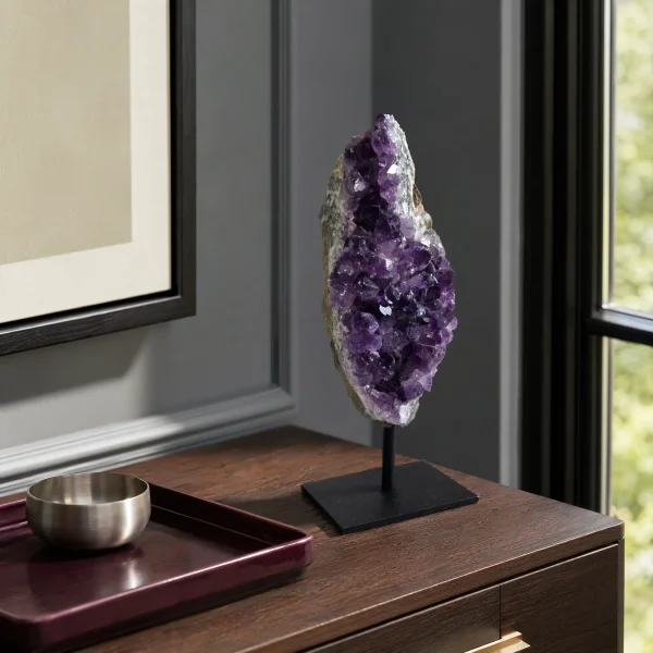 Amethyst cluster on a black metal stand displayed on a wooden cabinet near a window.