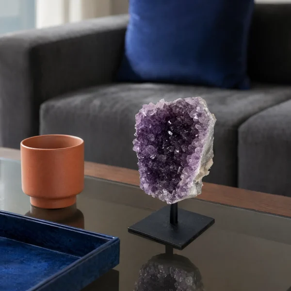 Amethyst cluster on a black metal stand displayed on a glass coffee table beside a cup and blue