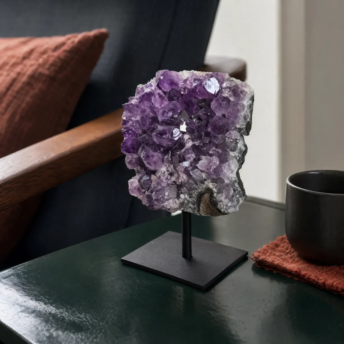 Amethyst cluster on a black metal stand displayed on a dark green side table beside a cup and chair.
