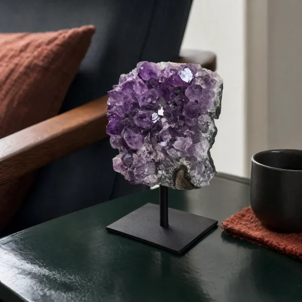 Amethyst cluster on a black metal stand displayed on a dark green side table beside a cup and chair.