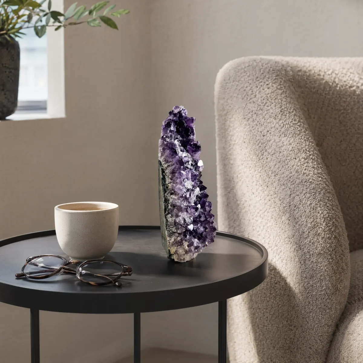 Small amethyst geode displayed on a black side table beside a cup, glasses, and an armchair.